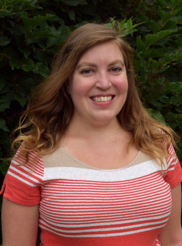 Alison Moffat, a white woman with shoulder length light brown hair, is standing in front of a leafy background. She is wearing a brightly colored pink and white top.