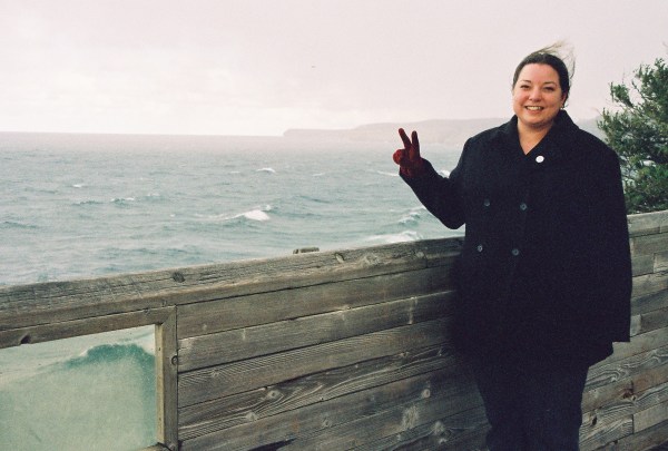 Leslie Mehle, a white woman with dark hair, is standing near a lake and showing the peace sign with her left hand.